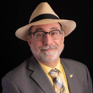 Headshot of Scott Lissner, wearing panama fedora hat, glasses, and a grey suit and 