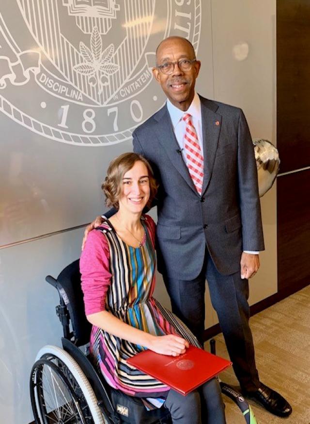 Jen Schlegel sits in her wheelchair with the Award certificate in a red leather case President Drake stands to her left with his hand resting lightly on her shoulder.